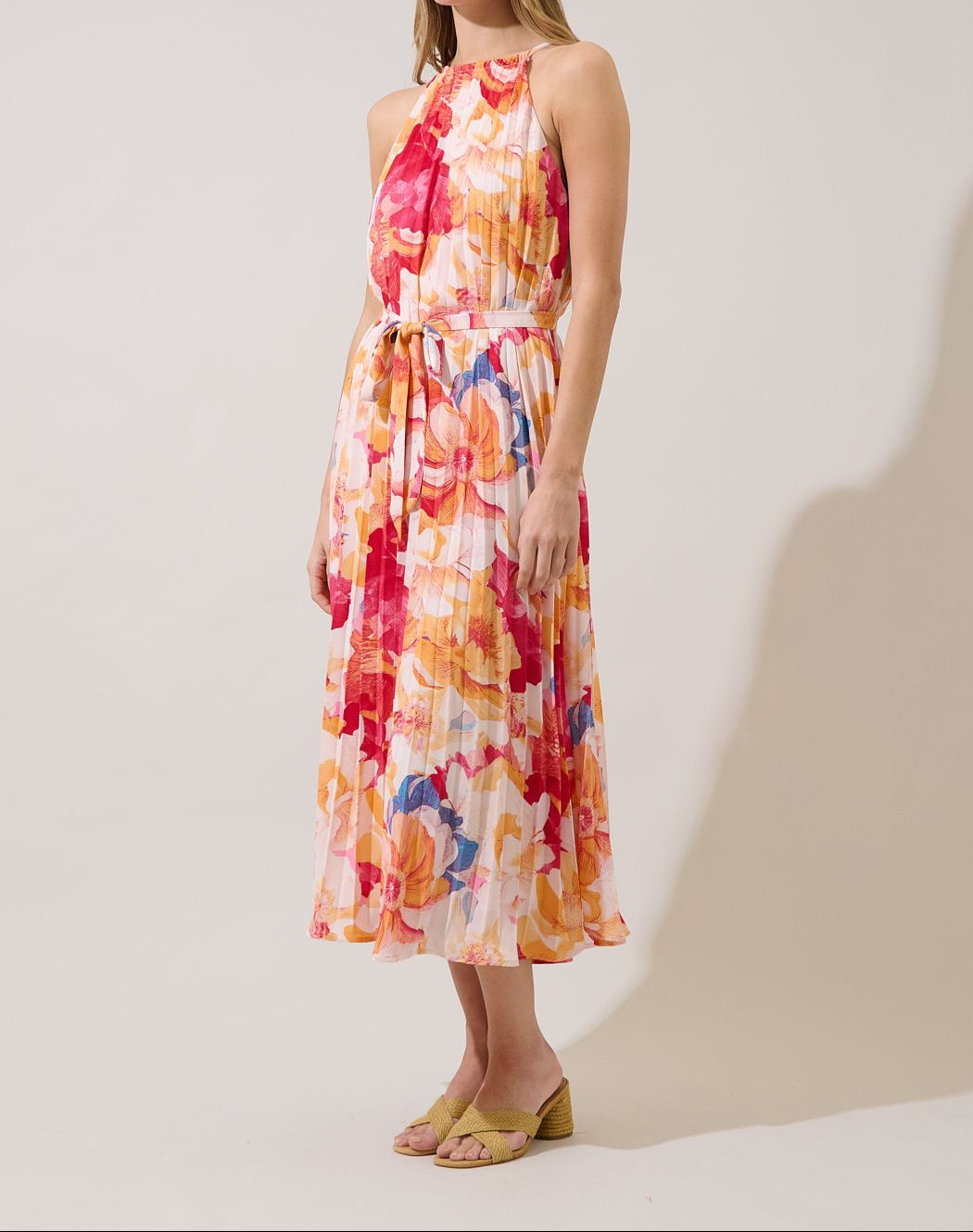Three-quarter view of coral floral pleated halter midi dress with a waist-defining self-tie belt