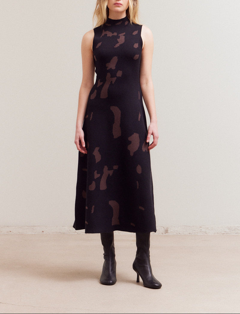 A model wearing a sleeveless black abstract patterned fitted  knit maxi dress with a high mock neckline.