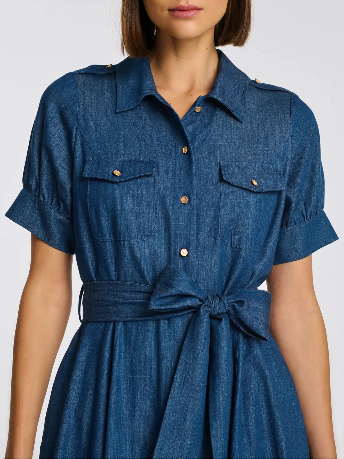 Close-up blue denim shirt dress with gold buttons, chest flap pockets, and tie waist